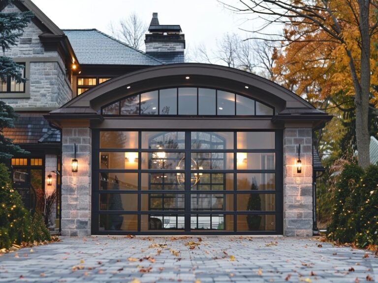 Elegant house with a stone exterior and large, arched glass garage door, illuminated by warm lights, set on a cobblestone driveway with autumn leaves.